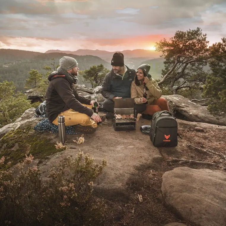 Ein Gruppe Personen sitzt auf einem Felsen im Wal. Im Hintergrund geht die Sonne unter. Die Personen grillen leckeres Fleisch auf einem Fennek Grill, daneben steht eine Tasche.