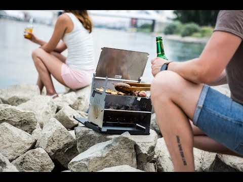 Mann baut den FENNEK Grill am See auf, befüllt ihn mit Kohle und zündet ihn an. Belegt den Rost mit Grillgut und grillt.