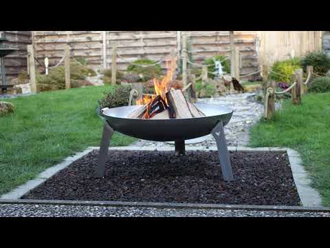 Die FENNEK Feuerschale 3Pod Edelstahl steht in einem kleinen Garten auf einem Kiesbett, darin brennt ein schönes Lagerfeuer.
