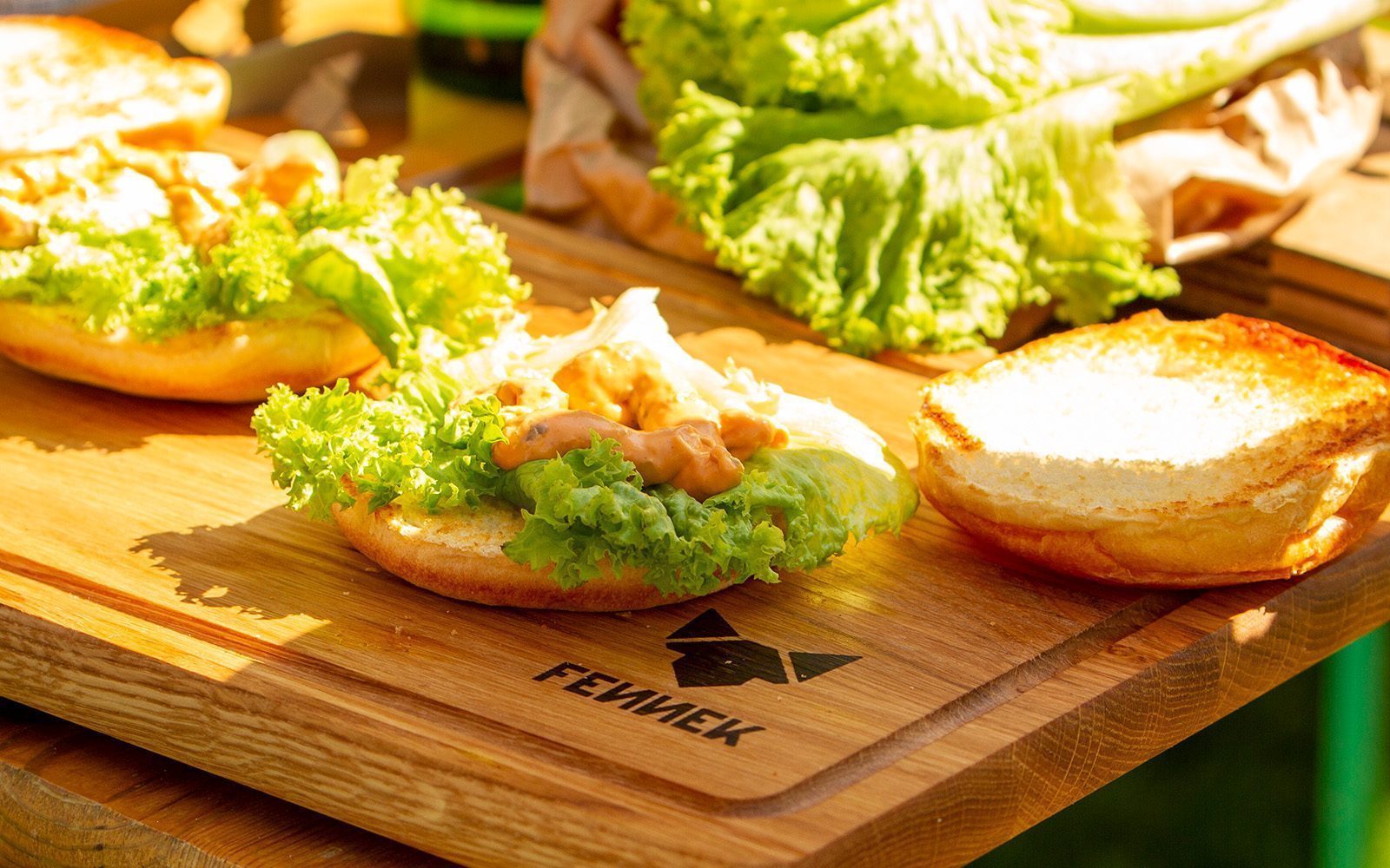 Brötchen für Hamburger mit Salatblättern und Sauce liegen auf einem Holz-Schneidebrett bei sonnigem Wetter.