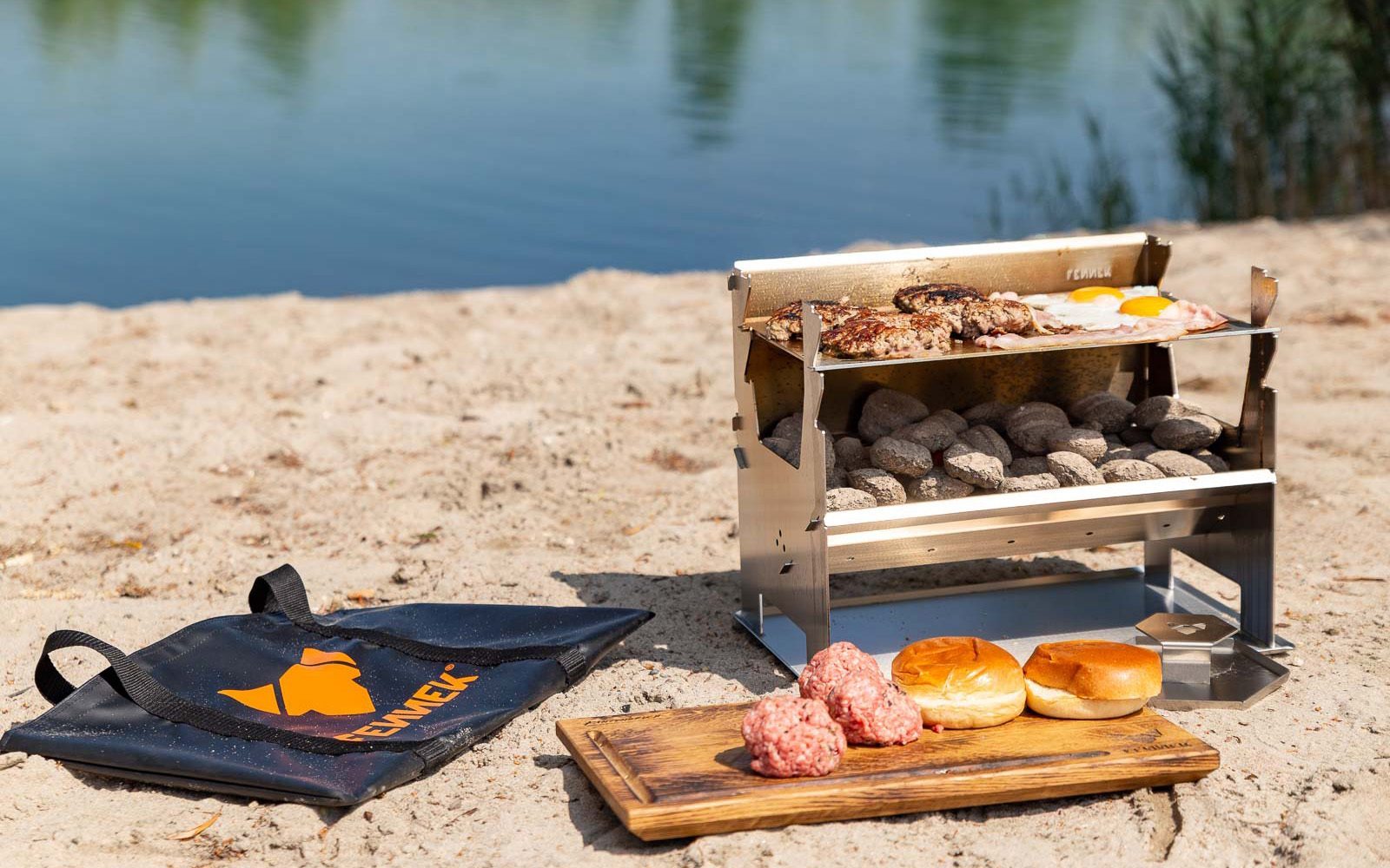 FENNEK 2.0 steht mir fast fertigem Grillgut auf der Plancha auf dem Strand. Vornedran liegt ein Schneidebrett auf dem Burgerpatties und Buns liegen. Die Tasche liegt daneben. Im HIntergrund ist ein See zu sehen.