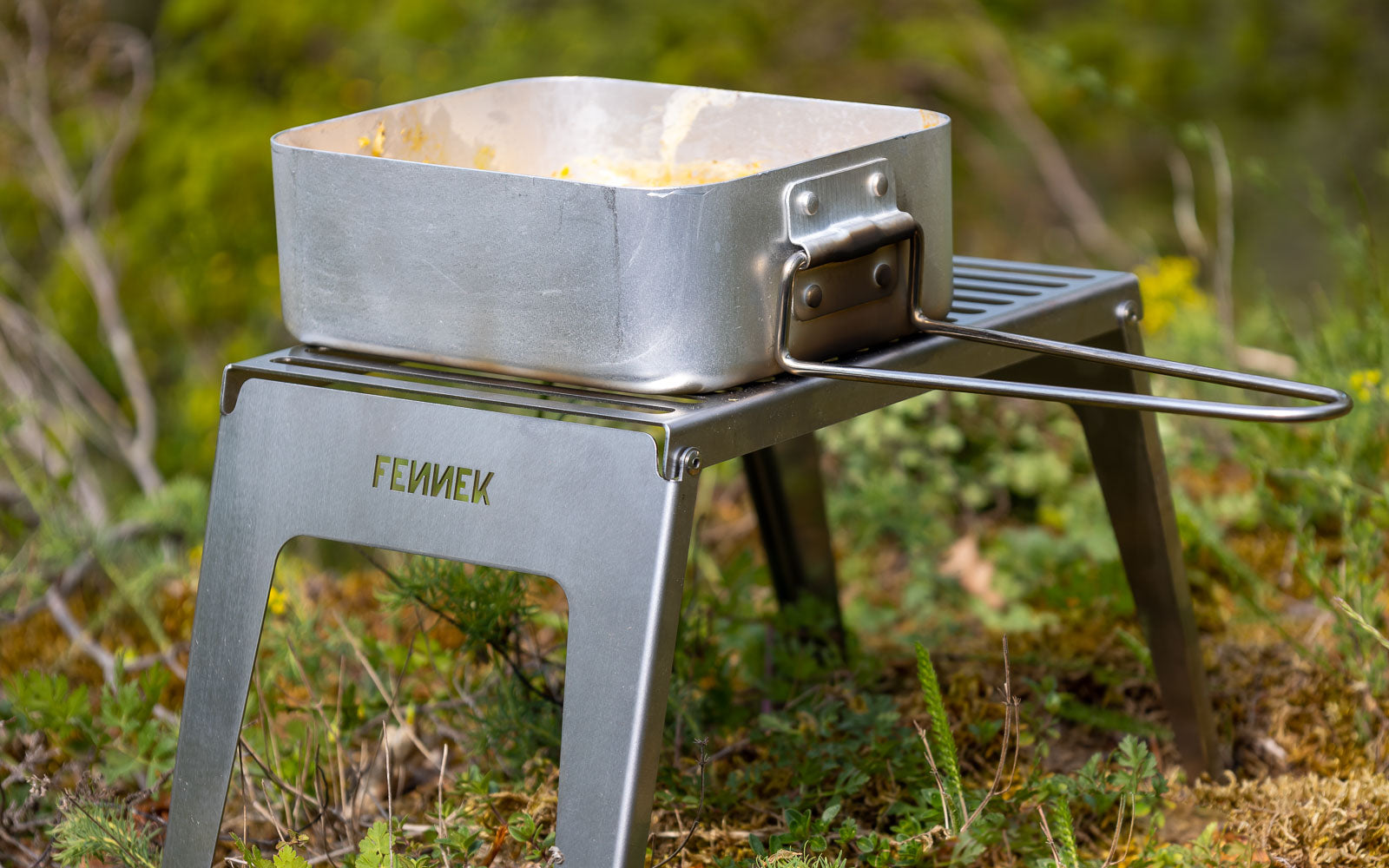 Der FENNEK Klapprost steht auf einer Wiese. Er fungiert als kleiner Tisch, weil eine Dose auf ihm platziert ist.