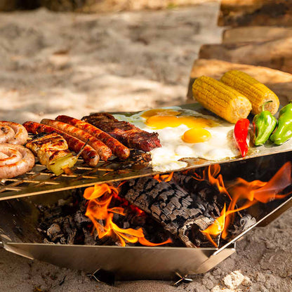 Bratwürste, Spiegelei und buntes Gemüse werden auf der Grillrost und -platte über der Feuerschale Hexagon die im Sand steht zubereitet.