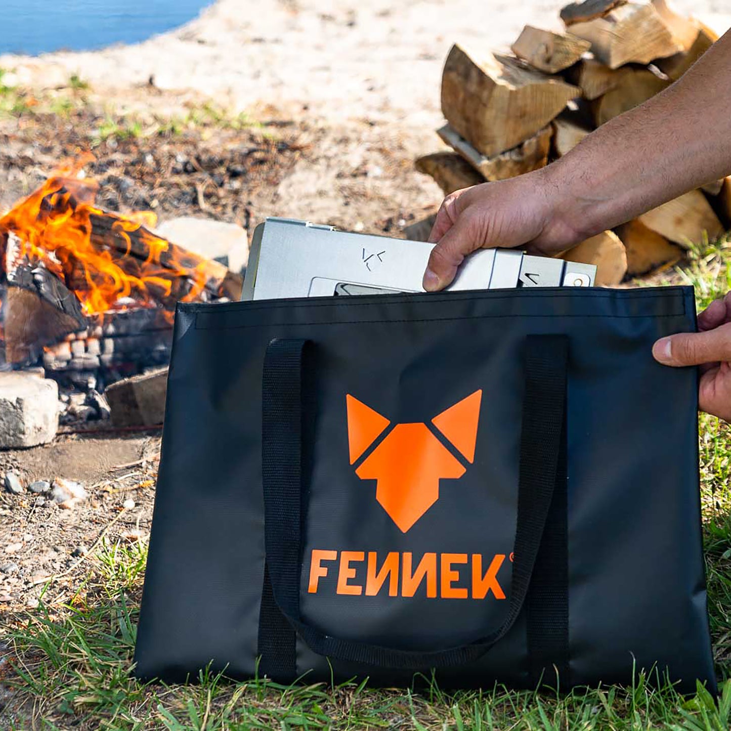 Einzelteile von Grillrost 4Fire werden aus der schwarzen Tarpaulin Henkeltasche mit FENNEK Logo-Aufdruck herausgezogen. Im Hintergrund brennt ein offenes Feuer.