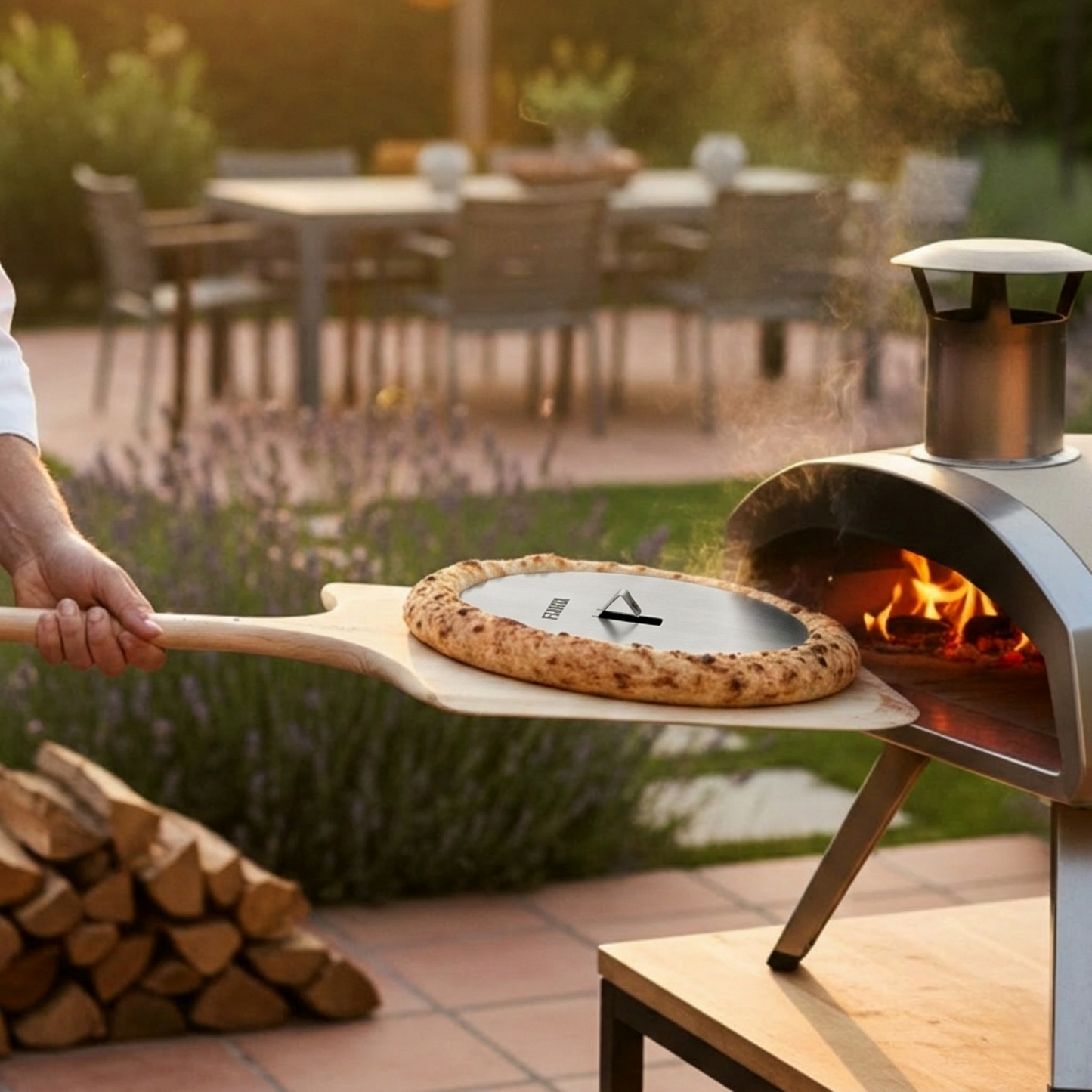 Szenebild zeigt einen kleinen Pizzaofen umgeben von einer schönen abendlichen Atmosphäre im Garten. Mit einem Pizzaschieber wird die Pizza aus dem Ofen geholt. Besonders auffällig ist der extra hohe, krosse und fluffige Rand der Pizza. Dieses Ergebnis wurde mit der FENNEK Blind Bake Edelstahlplatte erzielt, die genau dafür ausgelegt ist.