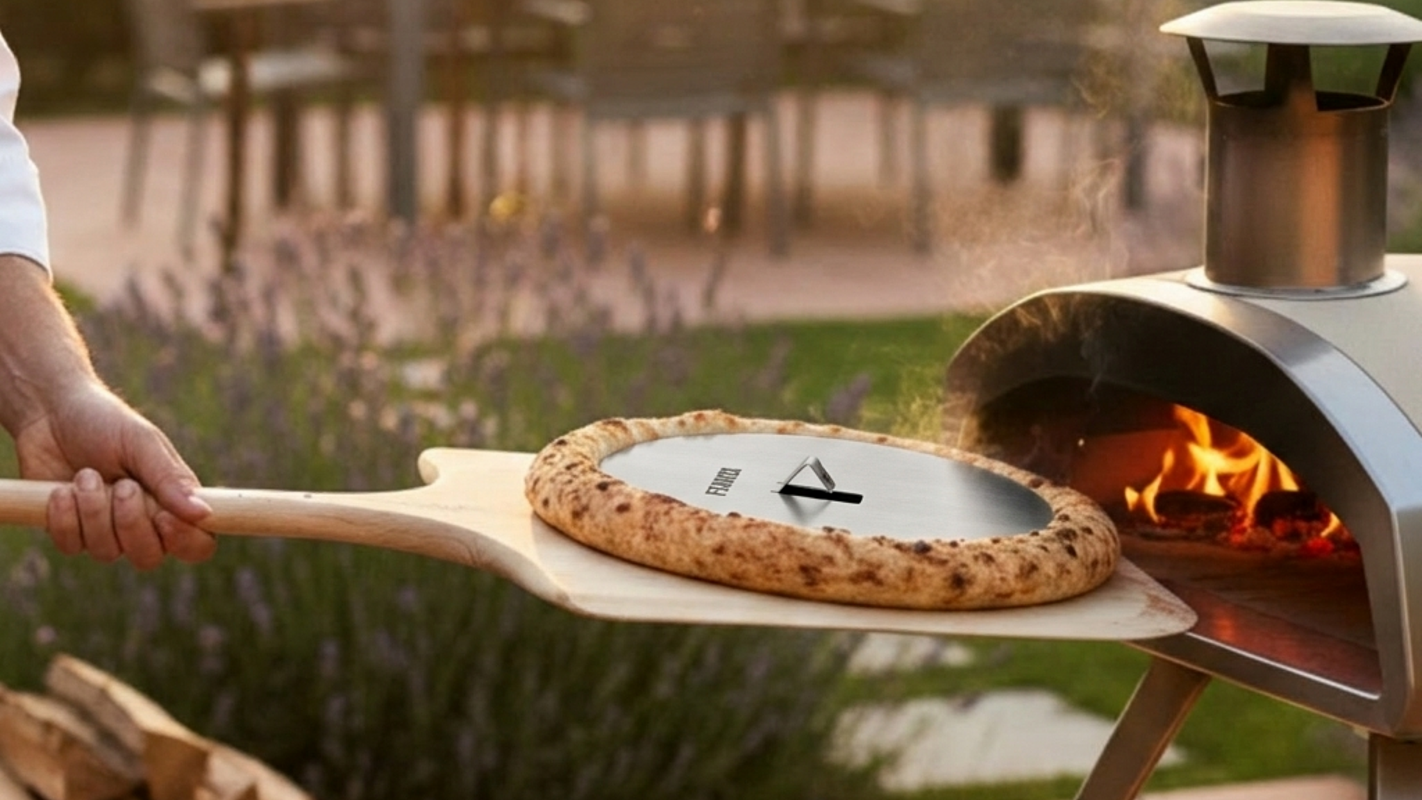 Szenebild zeigt einen kleinen Pizzaofen umgeben von einer schönen abendlichen Atmosphäre im Garten. Mit einem Pizzaschieber wird die Pizza aus dem Ofen geholt. Besonders auffällig ist der extra hohe, krosse und fluffige Rand der Pizza. Dieses Ergebnis wurde mit der FENNEK Blind Bake Edelstahlplatte erzielt, die genau dafür ausgelegt ist.