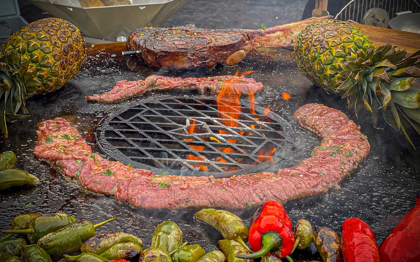 Szenebild FENNEK Feuerplatte - 96 cm inkl. optionalem Zubehör Grillrost. Auf der Feuerplatte werden ein Skirt-Steak, ein Tomahawk-Steak, 2 Ananas und diverse Paprika gegrillt. Aus der Mitte lodern Flammen durch den Grillrost. 