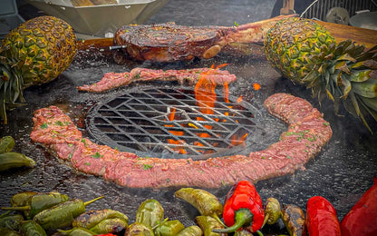 Szenebild FENNEK Feuerplatte - 96 cm inkl. optionalem Zubehör Grillrost. Auf der Feuerplatte werden ein Skirt-Steak, ein Tomahawk-Steak, 2 Ananas und diverse Paprika gegrillt. Aus der Mitte lodern Flammen durch den Grillrost. 