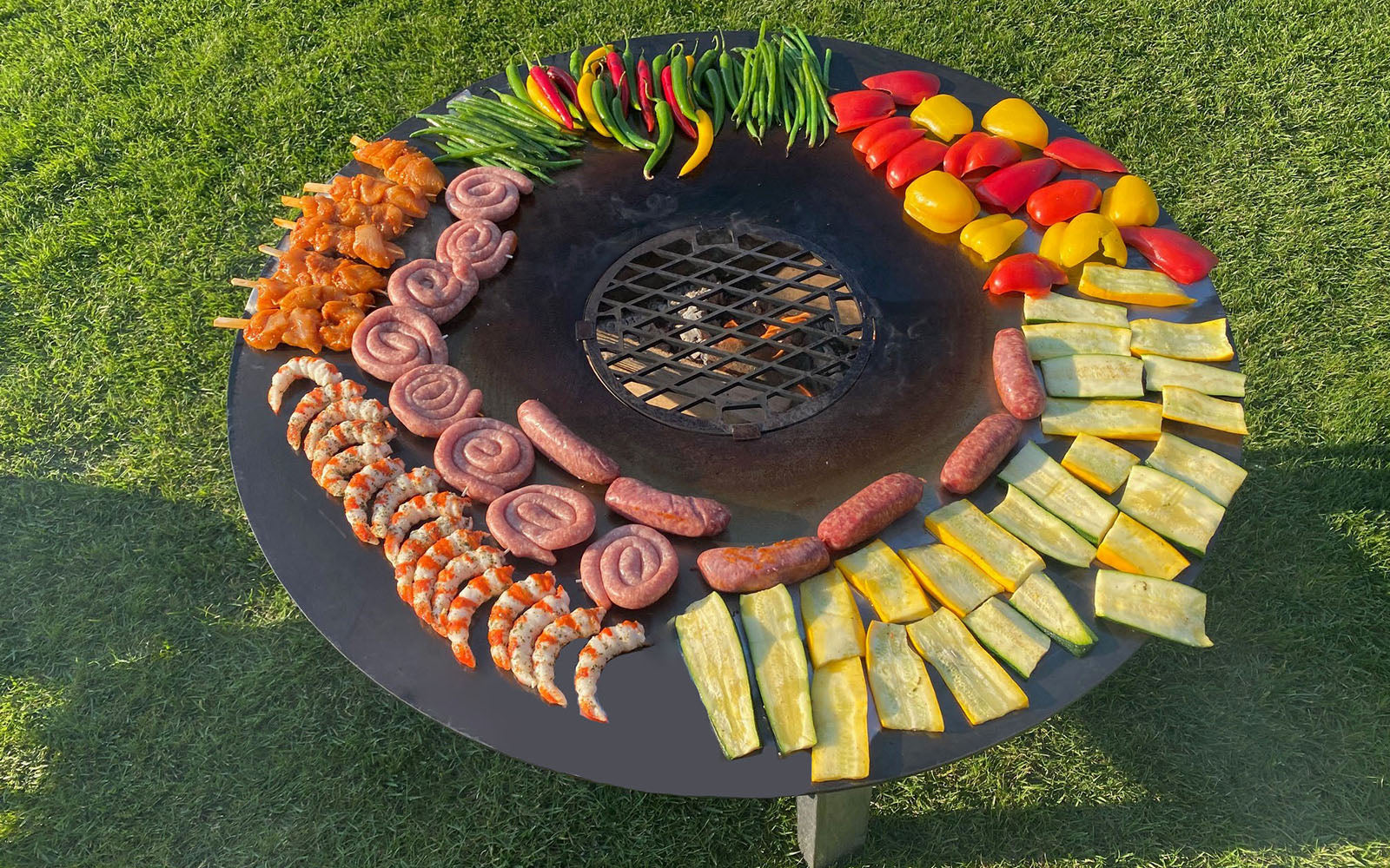 Szenebild der FENNEK Feuerplatte - 96 cm. Auf der Feuerplatte werden Bratwürste, Shrimps, Hähnchenspieße, Zucchinischeiben, Paprikascheiben und Peperoni gegrillt.