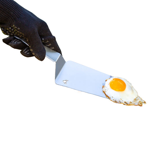 Schwarzer Grillhandschuh hält Edelstahl BBQ-Wender auf dem ein Spiegelei liegt