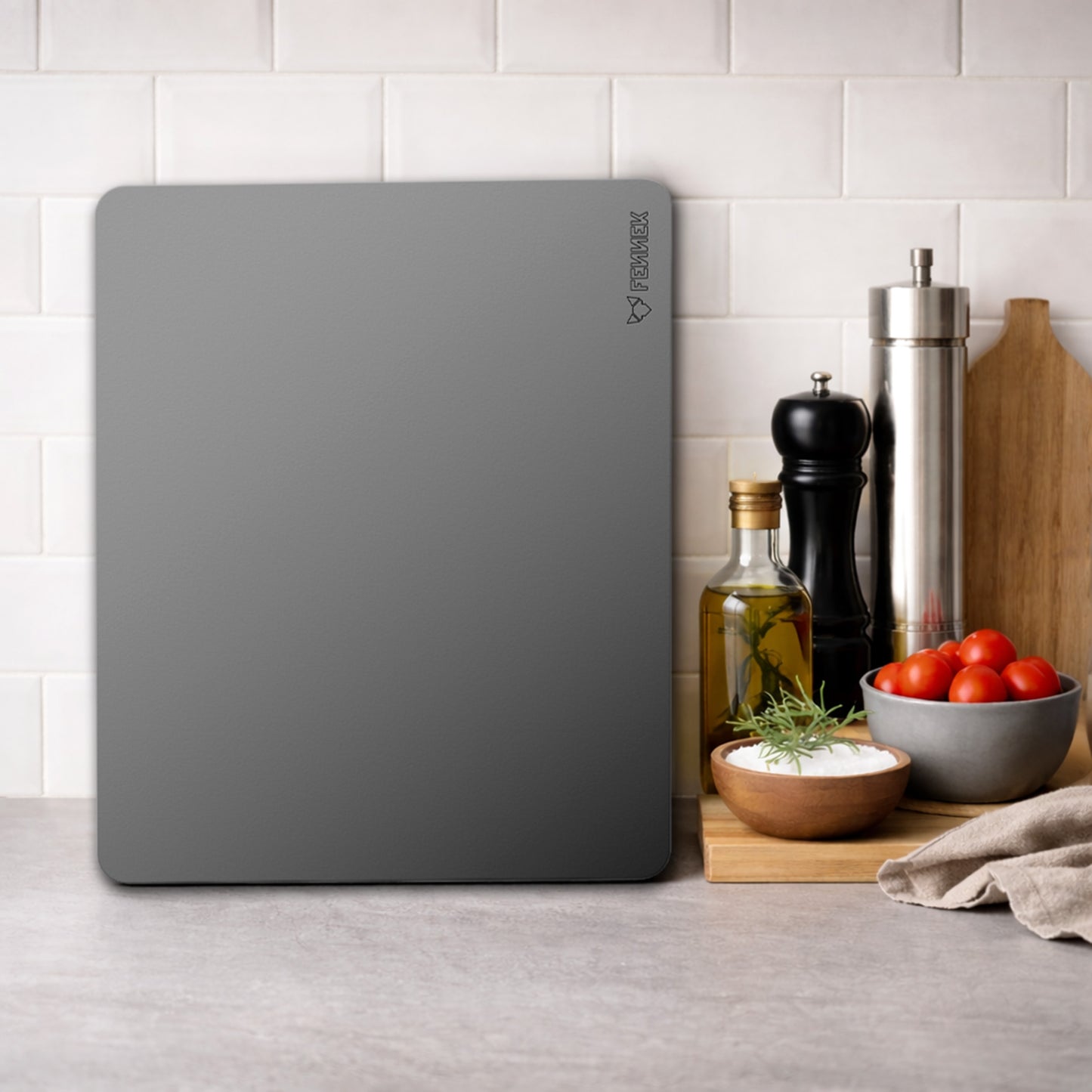 Der FENNEK Backstahl 40 x 30 cm steht senkrecht an einem Fliesenspiegel in einer Küche. Daneben steht eine Pfeffermühle, in Salzstreuer und eine Flasche mit Öl. Davor liegt ein kleines Holzschneidebrett mit Salz und Tomaten darauf. 