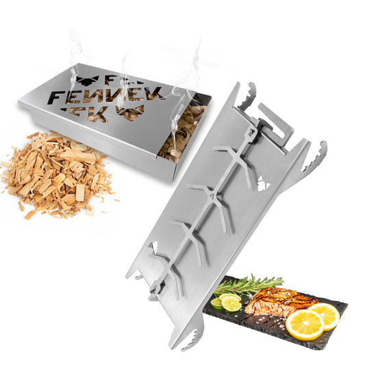 Links im Bild sieht man eine freigestellte FENNEK Räucherbox und ein paar Holzchips. Daneben der freigestellte FENNEK Flammlachshalter Shorty. Unten rechts im Bild sieht man einen fertig gegrillten Lachs, welcher auf einer Schiefertafel präsentiert wird. Garniert wird er mit Zitronen, Rosmarin und Limetten.