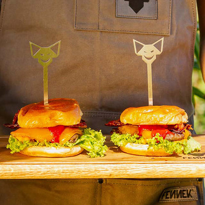 Edelstahl Burgerspieße mit FENNEK-Kopf-Griff stecken in zwei saftigen Cheeseburgern mit gebratenem Bacon und Salat, auf einem Schneidebrett aus Holz, gehalten von einer Person mit FENNEK Grillschürze.