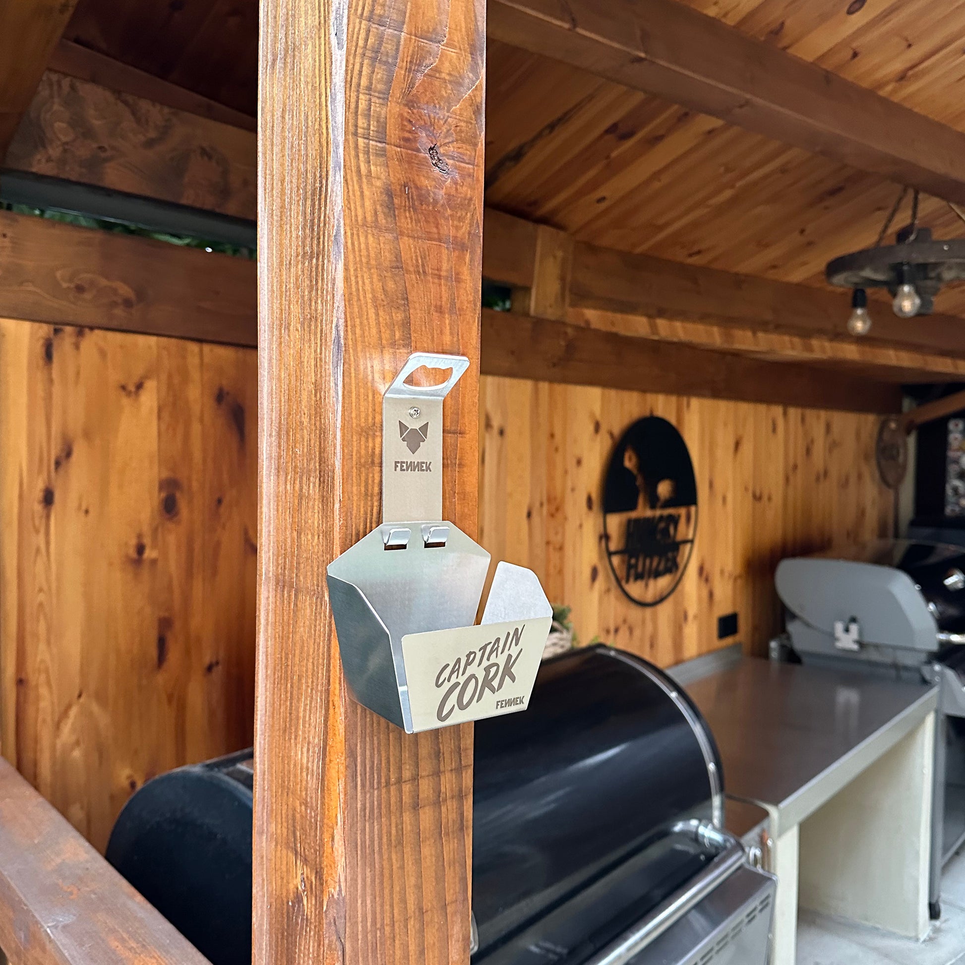 Der FENNEK Flaschenöffner Captain Cork mit Auffangkorb für bis zu 70 Kronkorken ist mit Schrauben an einer Grillecke angebracht. Im Hintergrund ist die Holzverkleidung der Ecke zu sehen, außerdem mehrere Grills. 