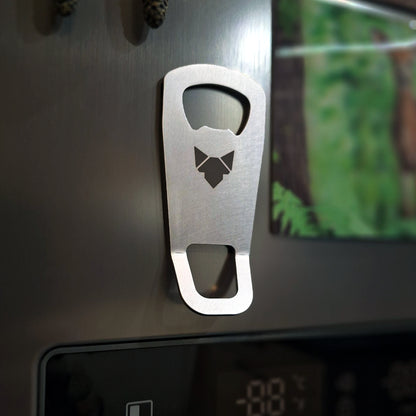 Der FENNEK Flaschenöffner Cpt. Cork - Magnetisch Mobil hängt dank seinem Magneten an einem grauen Edelstahl Kühlschrank. Rechts davon kann man ein Bild eines Rehs erkennen. Darunter ist die Temperatur-Anzeige des Kühlschranks zu erahnen. 