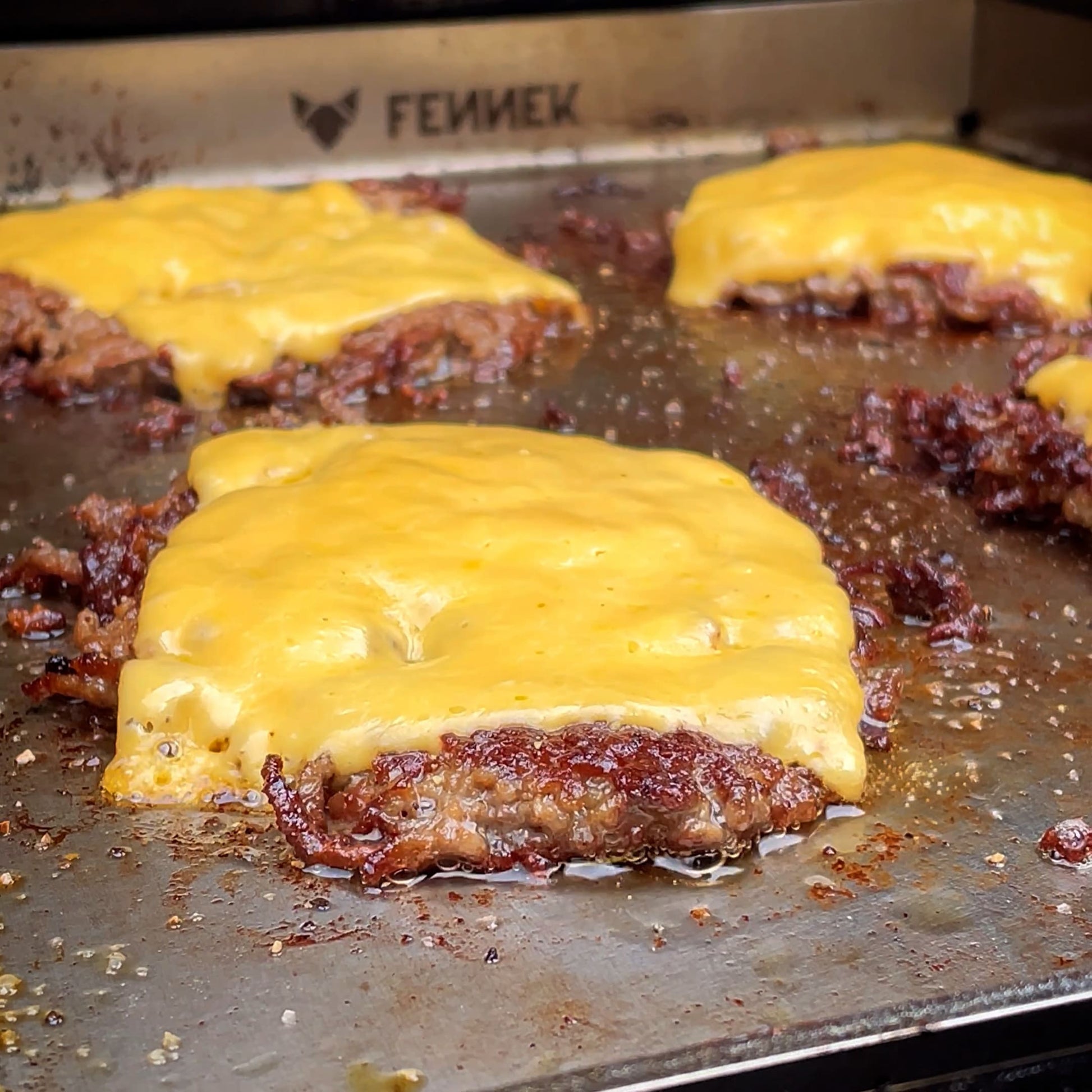 FENNEK Plancha 30 x 40 im Gasgrill. Man sieht Smashed Burger am braten, welche mit einer Scheibe Käse überbacken sind. Hinten in der Plancha ist das FENNEK Logo eingraviert.
