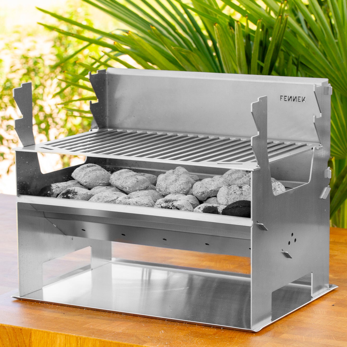 Fennek Holzkohlegrill 2.0 aus Edelstahl aufgebaut und mit brennender Kohle darin auf einem Bodenblech das auf einem Holztisch im Garten steht.