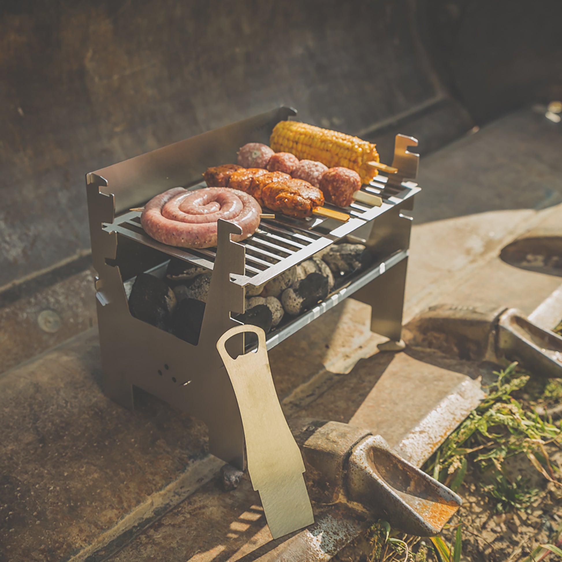 Mobiler Holzkohlegrill im Einsatz, steht in einer Baggerschaufel und grillt bei sonnigem Wetter verschiedenes Fleisch und einen Maiskolben.