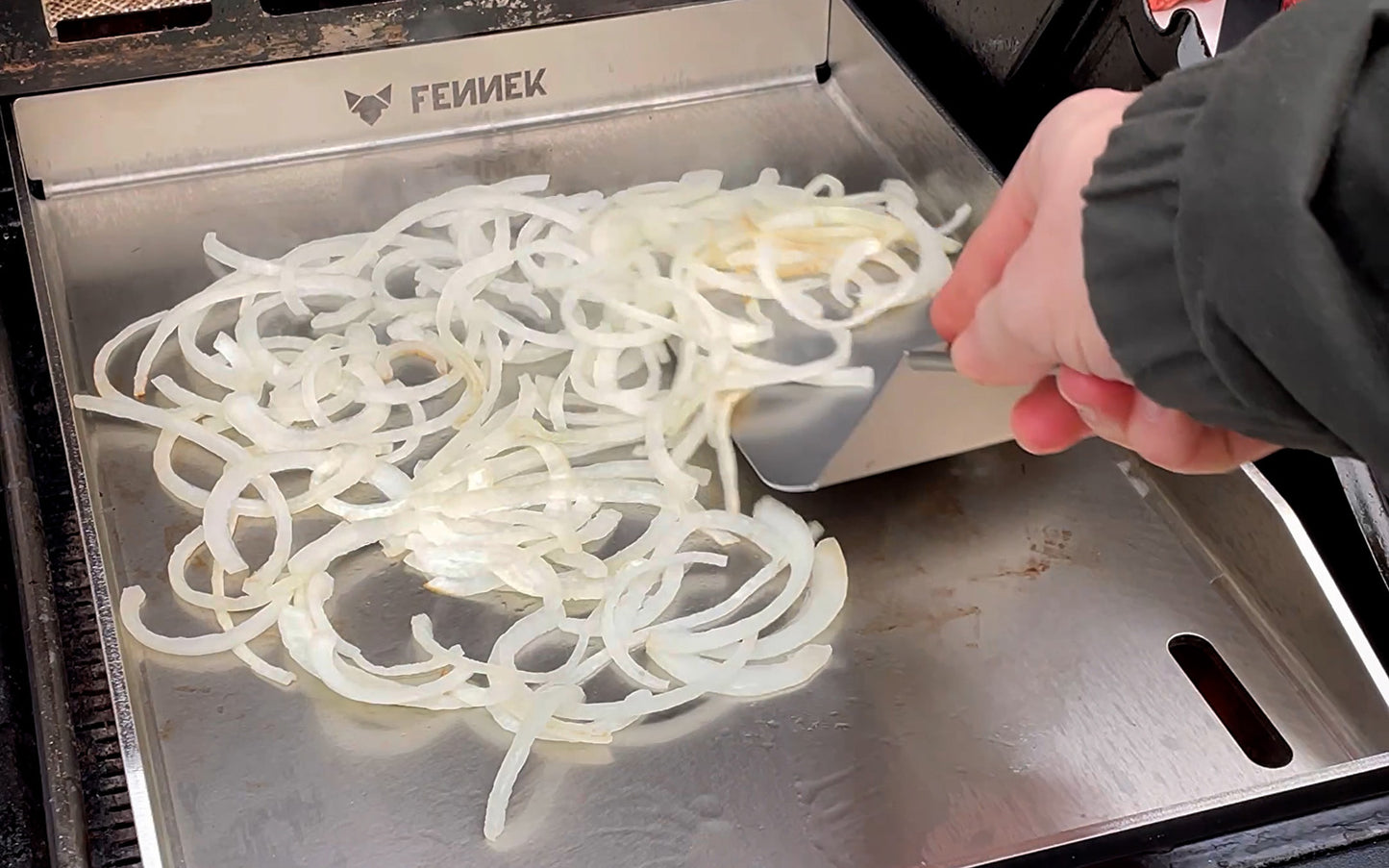 FENNEK Plancha 30 x 40 im Gasgrill. Es werden Zwibeln zubereitet und mit dem FENNEK BBQ Wender verteilt.