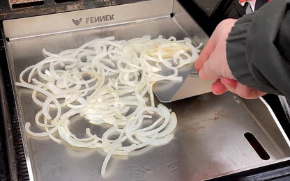 FENNEK Plancha 30 x 40 im Gasgrill. Es werden Zwibeln zubereitet und mit dem FENNEK BBQ Wender verteilt.