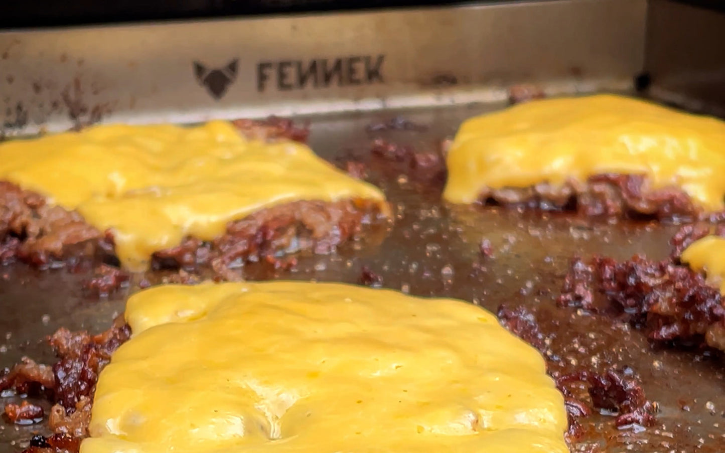 FENNEK Plancha 30 x 40 im Gasgrill. Man sieht Shmashed Burger am braten, welche mit einer Scheibe Käse überbacken sind. Hinten in der Plancha ist das FENNEK Logo eingraviert.