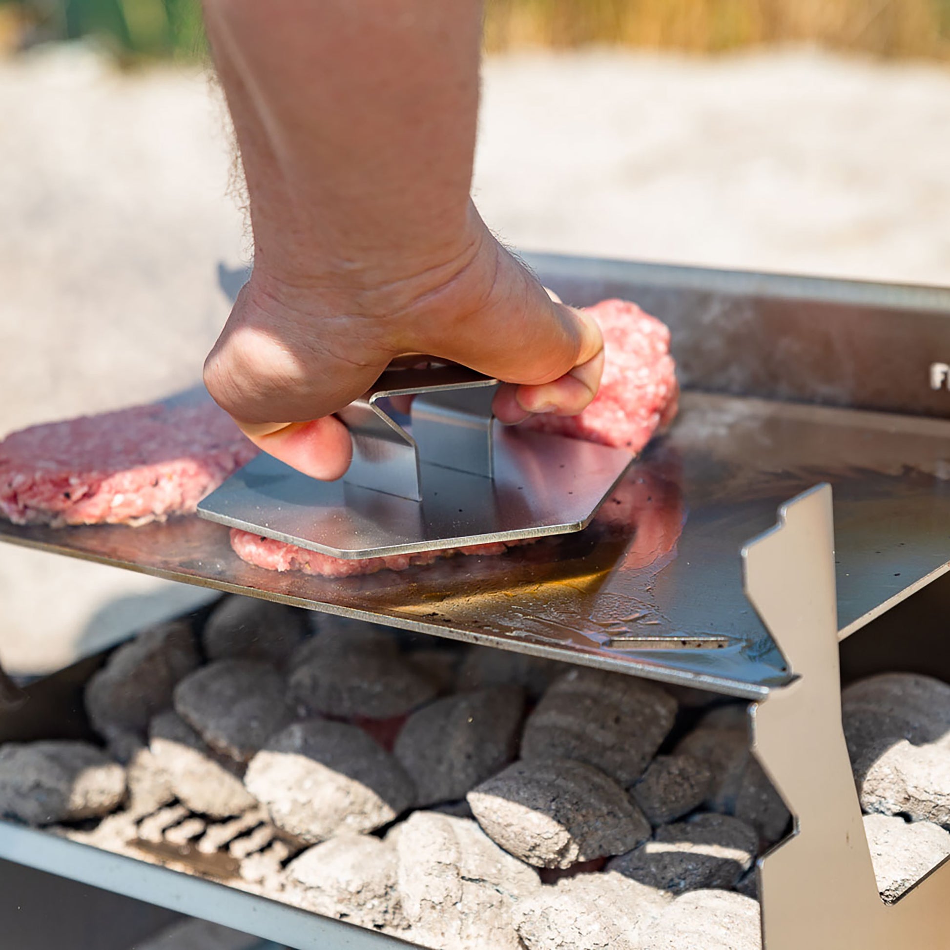 Eine Hand hält 6-eckige Hamburger Presse aus Edelstahl und presst einen Burger Patty auf der Plancha eines Holzkohlegrills.