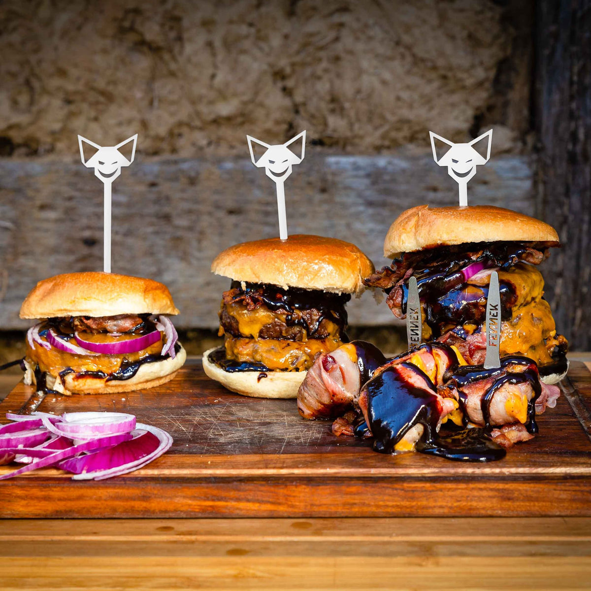 Ein Holzbrett mit drei aufsteigend großen Cheeseburgern und FENNEK Burgerspieß jeweils und davor Snickers-Riegel im Speckmantel gegrillt mit Fennek Pieker-Gabeln.