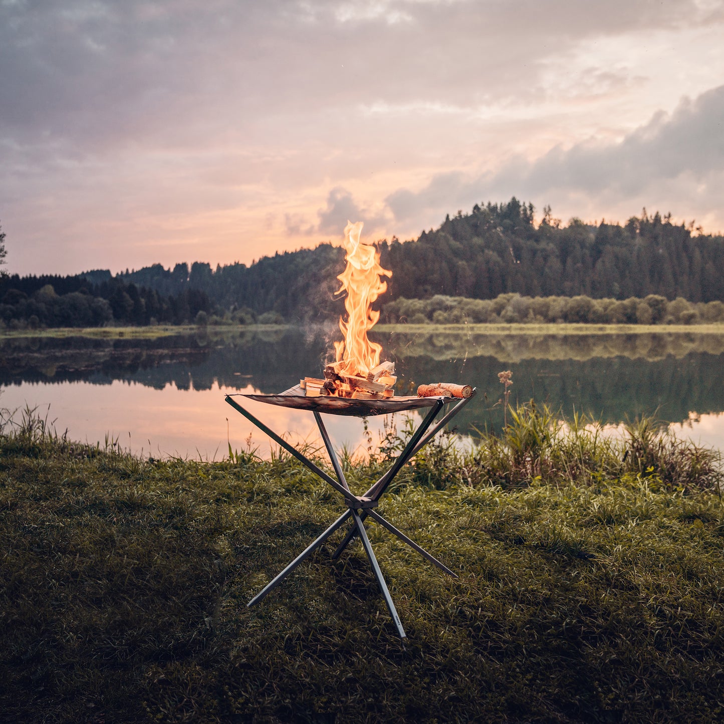 Das in der Natur aufgestellte Feuernetz im Einsatz mit brennendem Holz darauf und einer stimmungsvollen Aussicht auf ein stilles Gewässer mit Spiegelung der dichtbewachsenen Landschaft im Hintergrund.