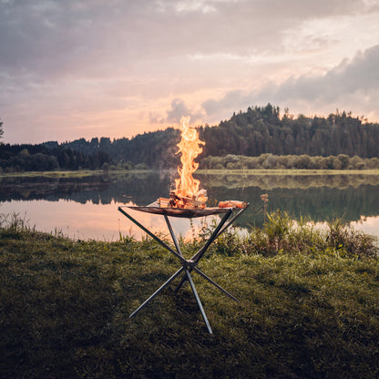 Das in der Natur aufgestellte Feuernetz im Einsatz mit brennendem Holz darauf und einer stimmungsvollen Aussicht auf ein stilles Gewässer mit Spiegelung der dichtbewachsenen Landschaft im Hintergrund.