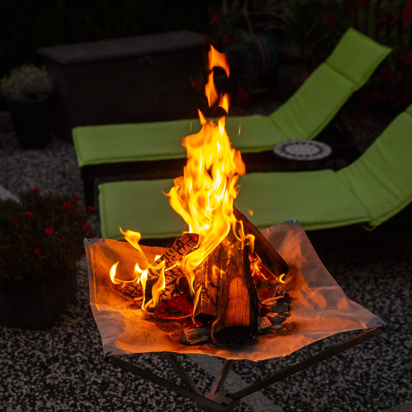 Brennendes Feuer im Feuernetz bei Dunkelheit, das im Garten steht und im Hintergrund zwei Sonnenliegen.