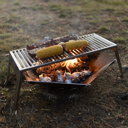 Der FENNEK Klapprost XL steht über der FENNEK Feuerschale Hexagon in welcher glühende Kohle und Kartoffel in Alufolie liegen Alles steht auf grünem Gras.