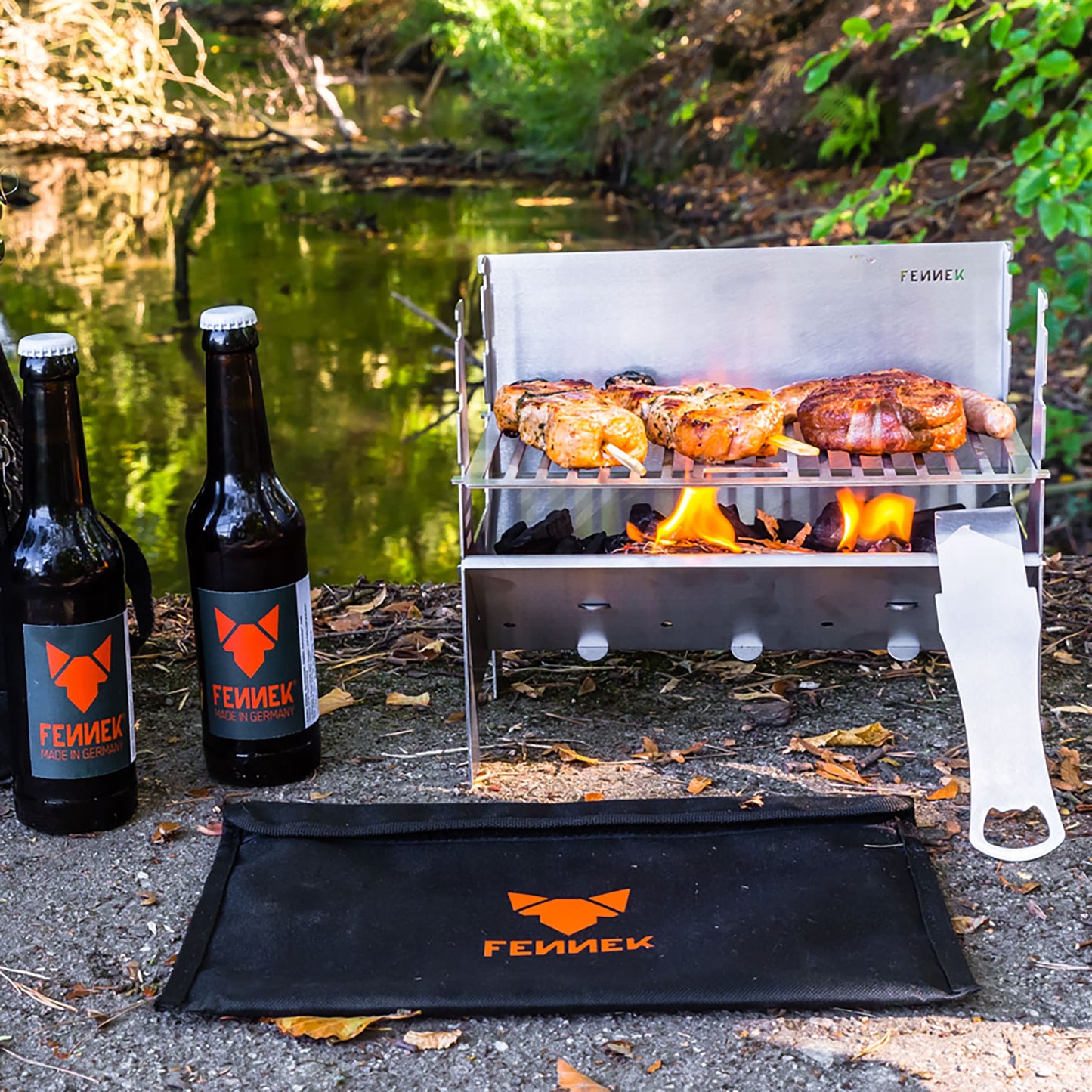 Im Freien aufgebauter Fennek Grill aus Edelstahl auf dem Grillfleisch über brennender Kohle gegrillt wird, daneben 2 Flaschen mit FENNEK-Etikett und vor dem Grill liegt die schwarze Nylon-Tasche mit FENNEK Logo-Aufdruck, unscharf im Hintergrund Gewässer und viel grüne Natur.