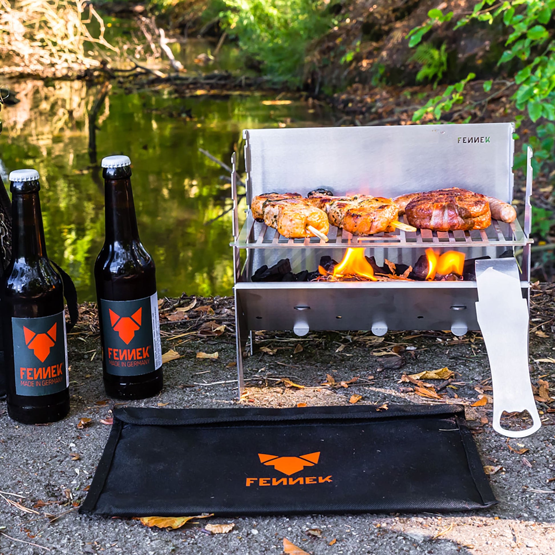 Im Freien aufgebauter Fennek Grill aus Edelstahl auf dem Grillfleisch über brennender Kohle gegrillt wird, daneben 2 Flaschen mit FENNEK-Etikett und vor dem Grill liegt die schwarze Nylon-Tasche mit FENNEK Logo-Aufdruck, unscharf im Hintergrund Gewässer und viel grüne Natur.