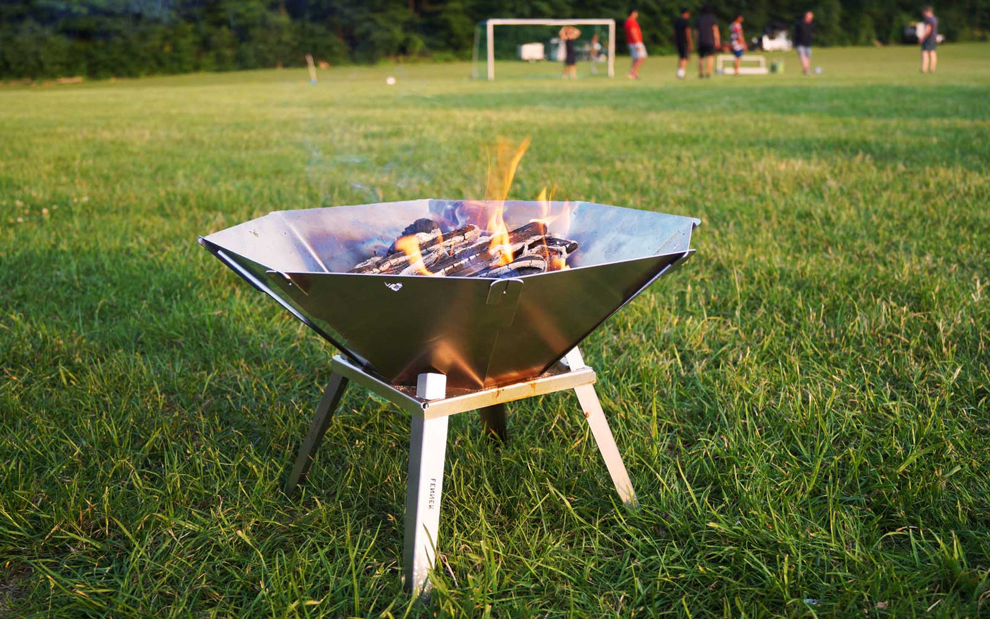 Holz brennt in einer Edelstahl Feuerschale steht mit 4 Standfüßen auf einer großen Wiese und im Hintergrund wird Fußball gespielt.