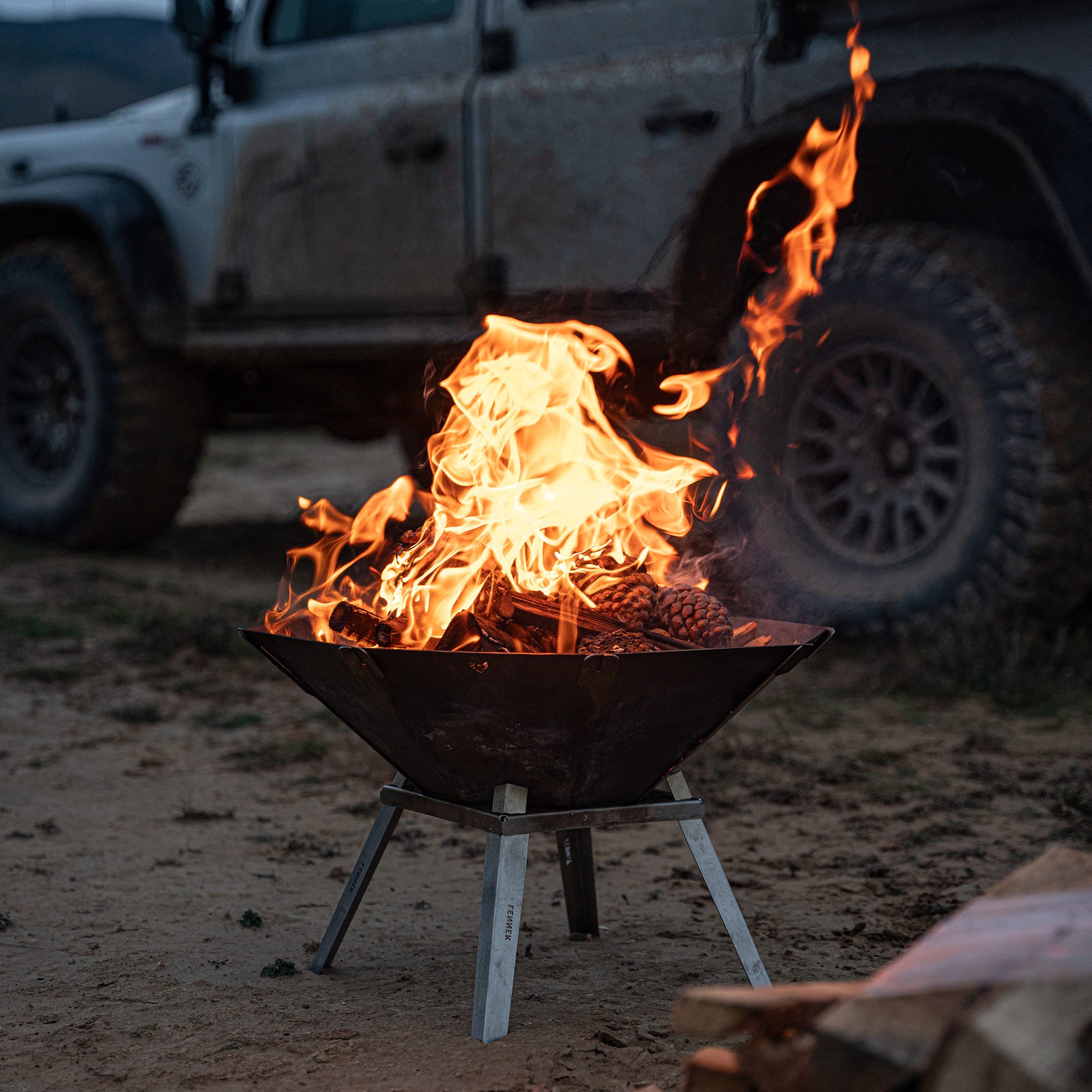 Die FENNEK Feuerschale Oktagon mit den optionalen Füßen steht vor einem Land Rover am Strand. Es ist Abendstimmung. Ein schönes Feuer lodert in der Feuerschale.