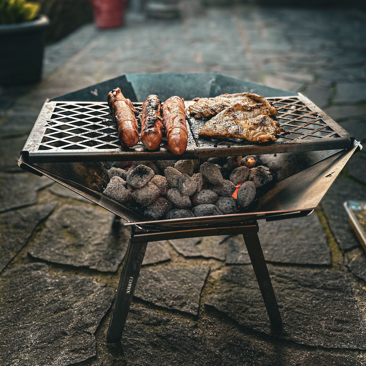 Die mobile, kompakte, steckbare Feuerschale Oktagon aus Edelstahl steht aufgebaut mit Füßen und Grillrost auf einer Steinterrasse. Es werden Berner Würstchen und Hähnchensteaks über glühender Glut gegrillt. 