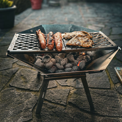 Die mobile, kompakte, steckbare Feuerschale Oktagon aus Edelstahl steht aufgebaut mit Füßen und Grillrost auf einer Steinterrasse. Es werden Berner Würstchen und Hähnchensteaks über glühender Glut gegrillt. 