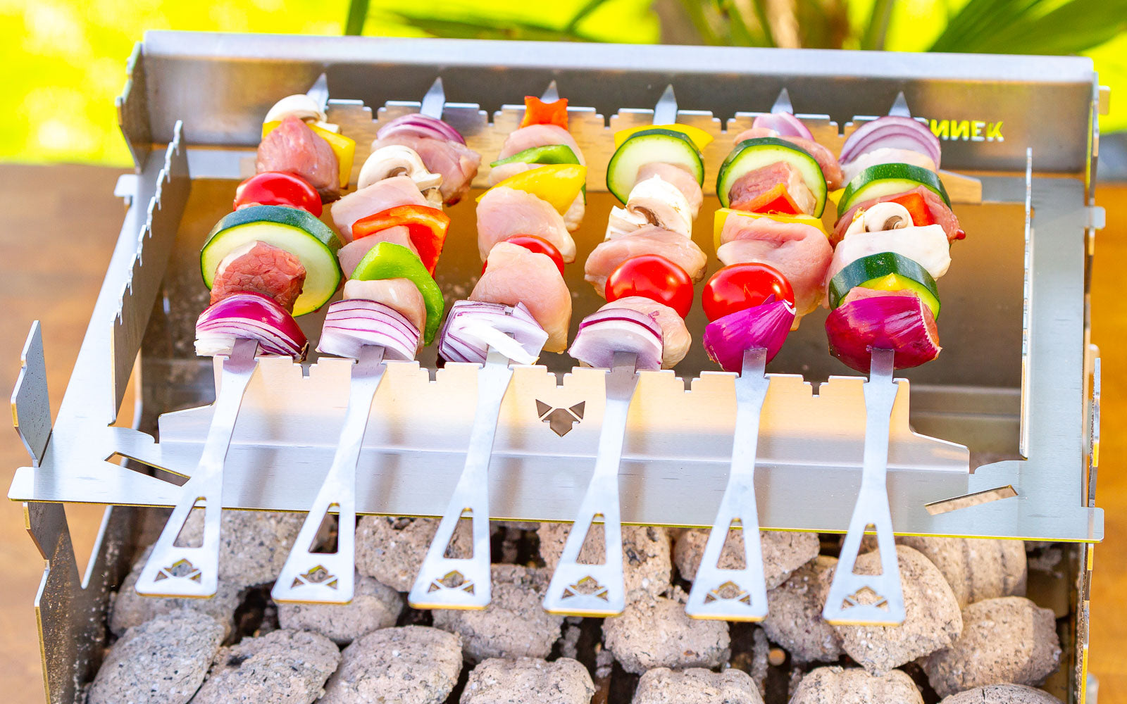Der FENNEK 2.0 mit Grillspießhalterung steht auf einem Tisch im Garten und auf ihm grillen sechs Häähnchenspieße.