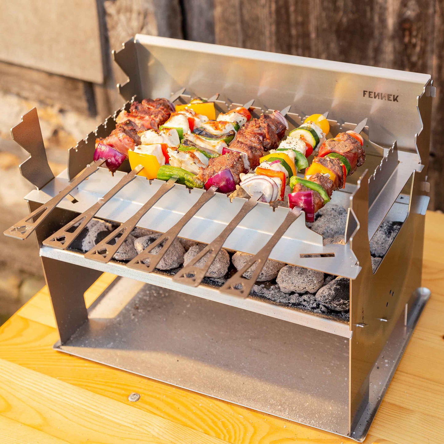 Bunt bestückte Grillspieße auf einem FENNEK Holzkohlegrill mit Spießhalterung und unter dem Grill ein Bodenblech das den Holztisch vor Schmutz schützt.
