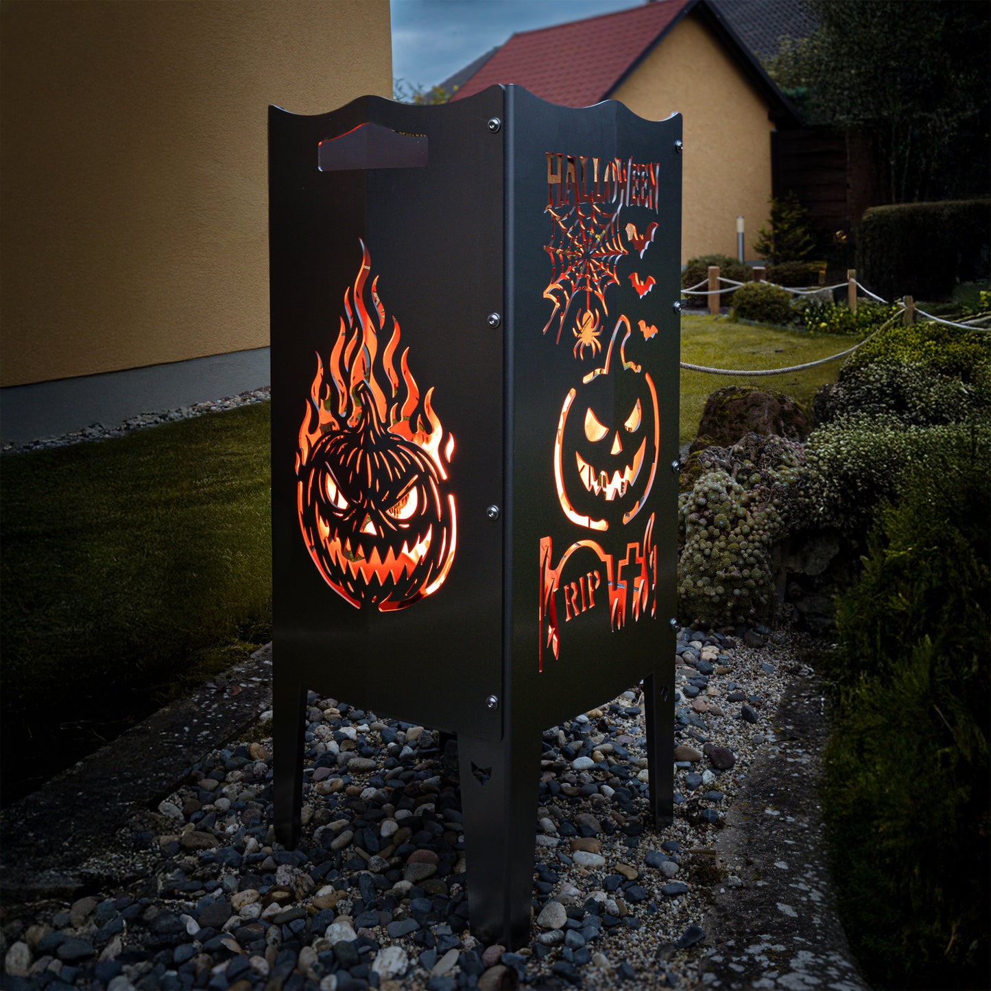 Die FENNEK Halloween Feuersäule lt. Edition steht in einem schönen Garten und wird Feuerholz von Innen beleuchtet. Der Feuerkorb kann aber auch mit einem LED-Strahler von Innen betrieben werden. Made in Germany.