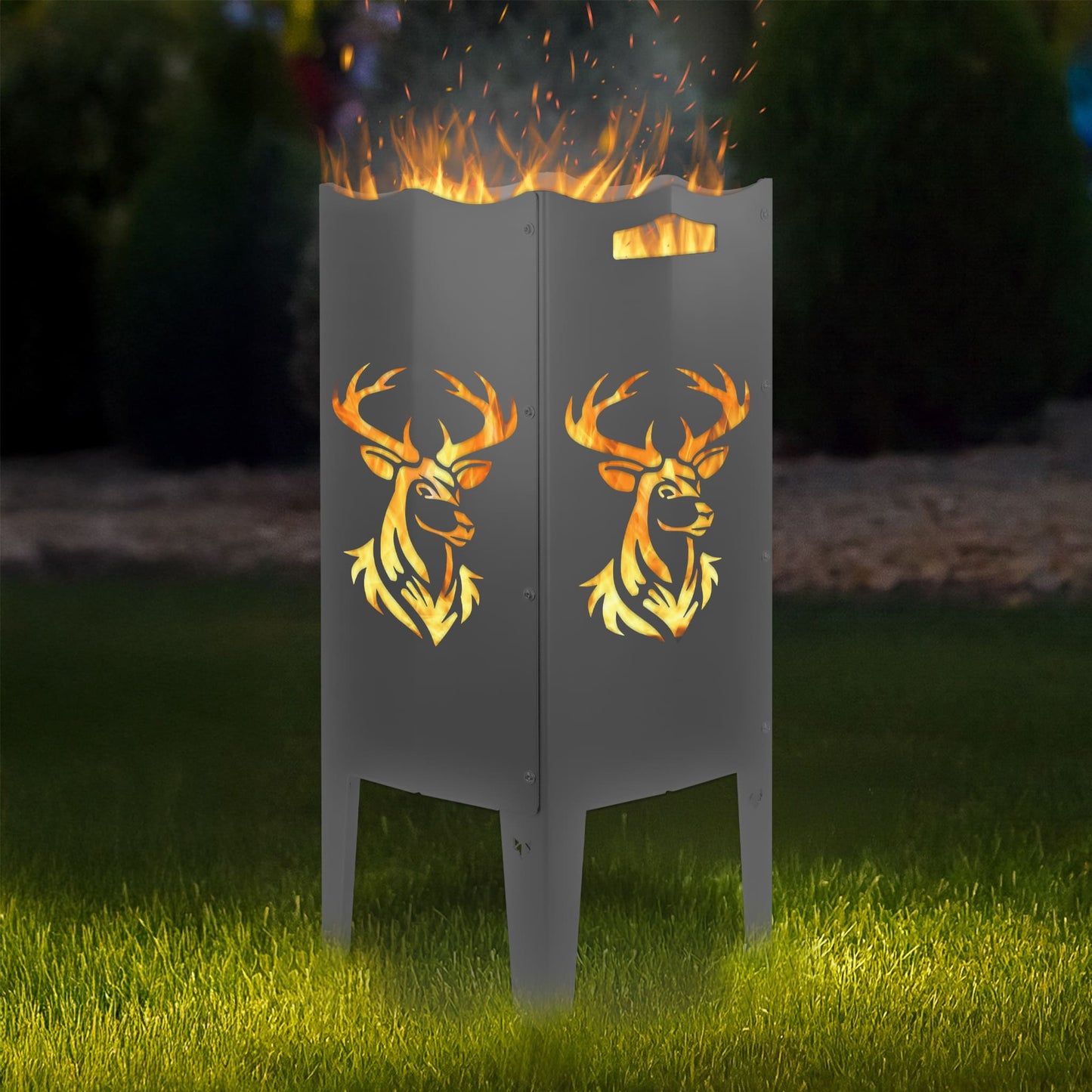 FENNEK Feuersäule Stahl mit dem Motiv Hirsch - in der Feuersäule brennt ein Feuer, man sieht das Motiv sehr schön leuchten in der Abenddämmerung in einem Garten. Die Feuersäule steht auf Gras.