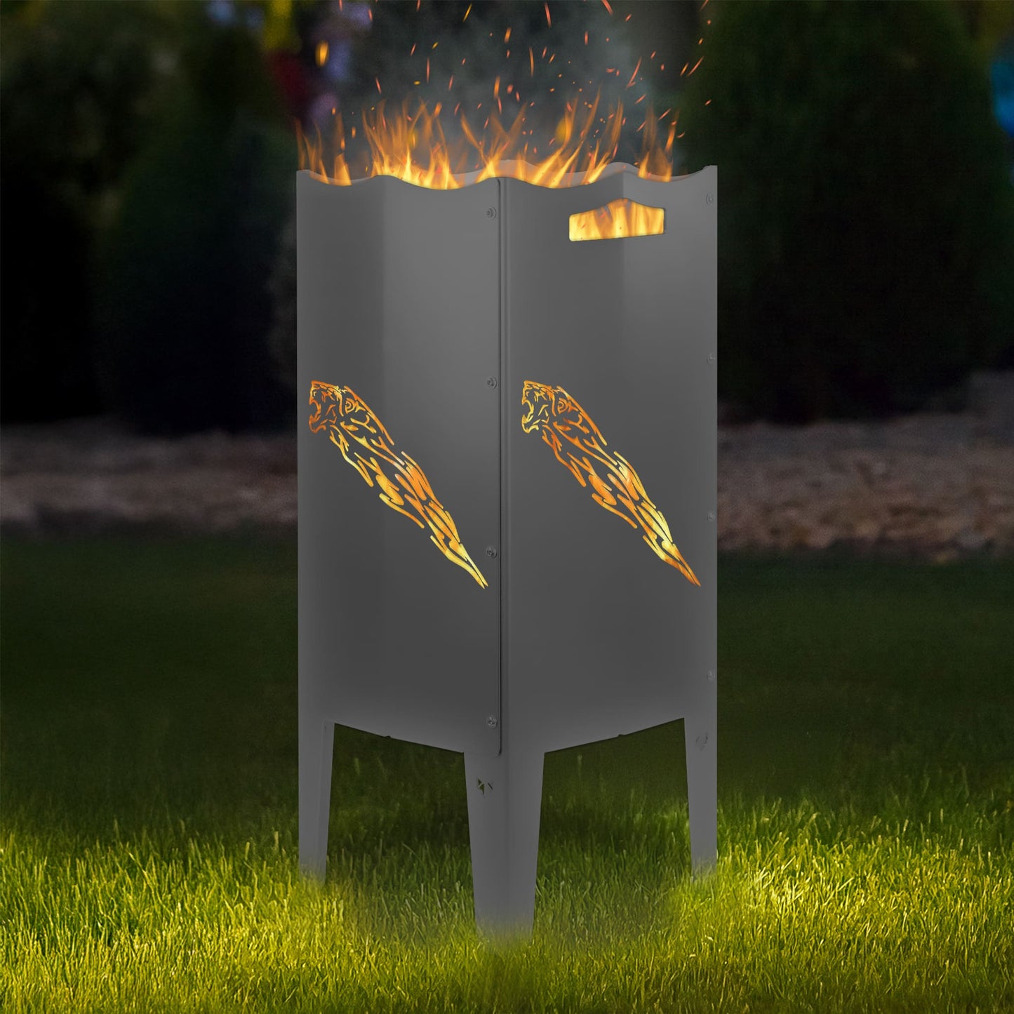 FENNEK Feuersäule Stahl mit dem Motiv Löwe mit Schweif - in der Feuersäule brennt ein Feuer, man sieht das Motiv sehr schön leuchten in der Abenddämmerung in einem Garten. Die Feuersäule steht auf Gras.