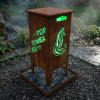 Die FENNEK Feuersäule aus Stahl in schönem Rost-Look steht in einem schönen Garten auf einem Kiesbett in ihrer Aschewanne aus Edelstahl. Die Aschewanne ist befüllt mit Kieselsteinen und dient somit nicht nur zum Schutz des Untergrunds, sondern auch als Deko-Element. Der passende Deckel aus Stahl schützt die Feuersäule vor Witterung. Natürlich Made in Germany!