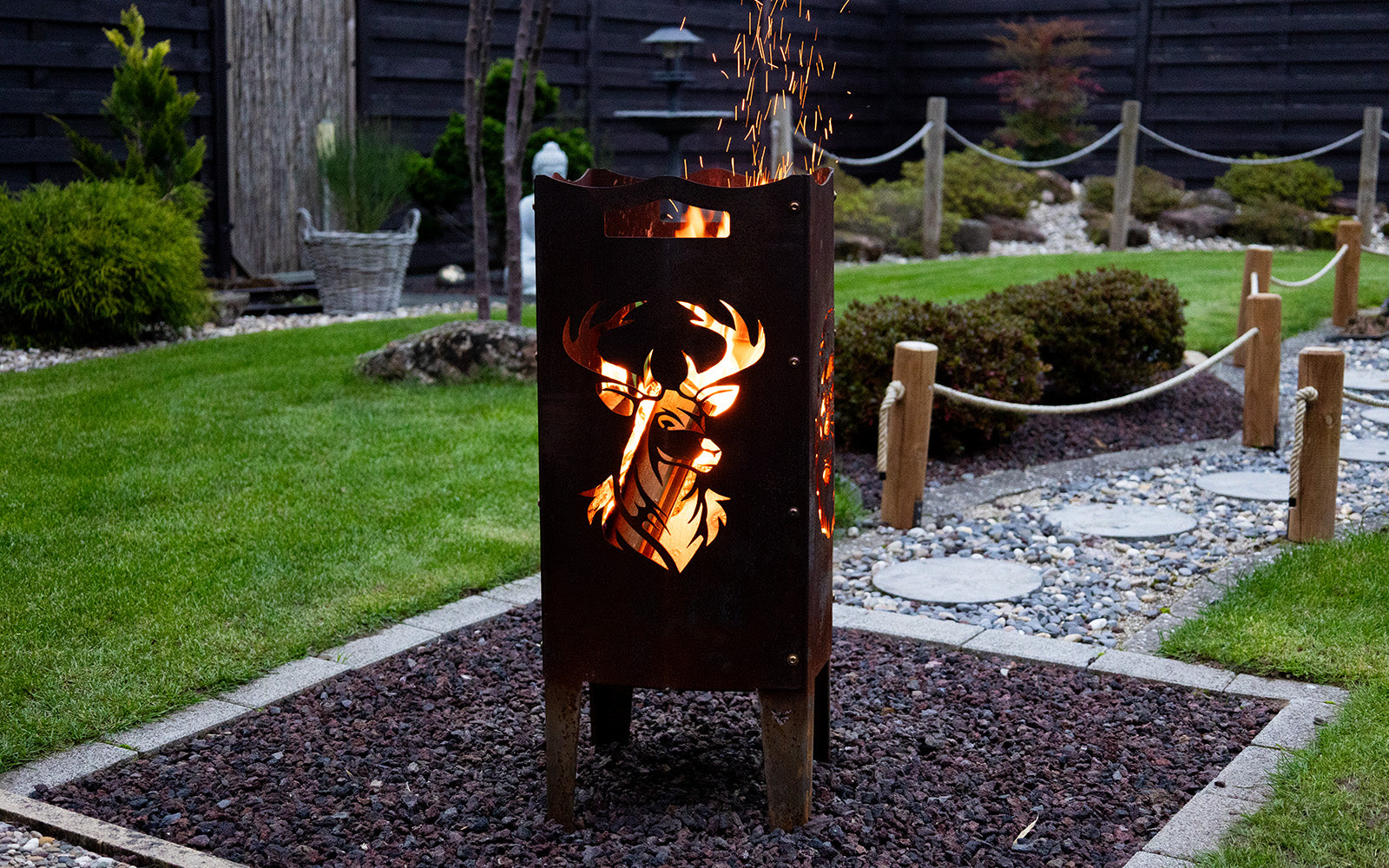 Feuersäule aus Stahl mit Hirsch Motiv steht in einem schönen leicht asiatisch angehauchten Garten. Die Feuersäule besitzt eine schöne Rostoptik und ein loderndes Feuer brennt darin, sodass der Hirsch sehr gut zur Geltung kommt.