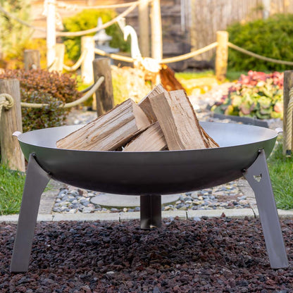 Die Feuerschale Stahl steht in einem Garten auf einem kleinen Kiesbett. In der Feuerschale liegt das Feuerholz bereit zum Entfachen.