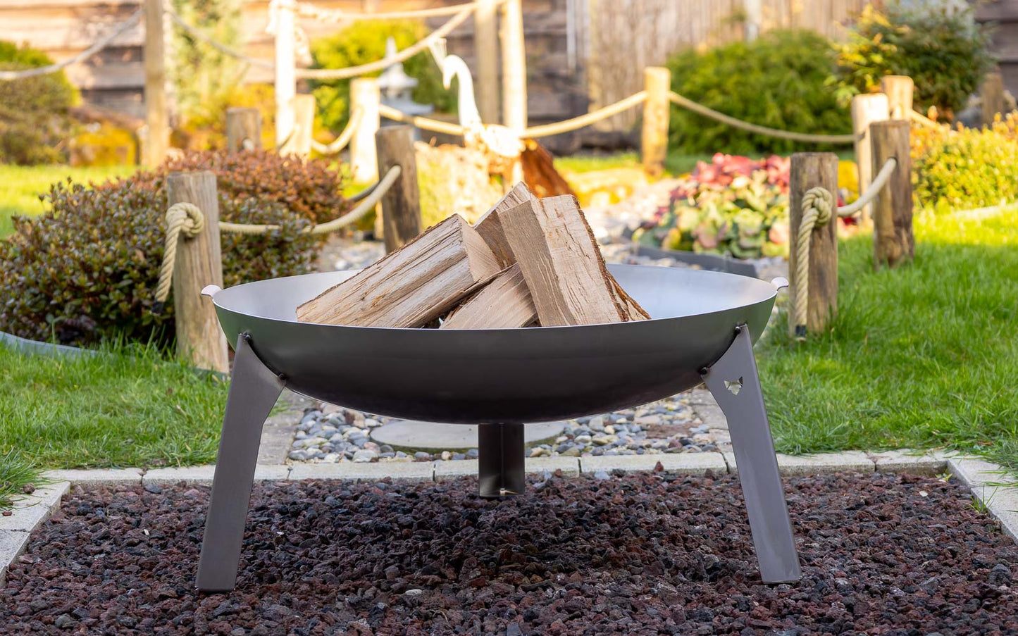 Die Feuerschale Stahl steht in einem Garten auf einem kleinen Kiesbett. In der Feuerschale liegt das Feuerholz bereit zum Entfachen.