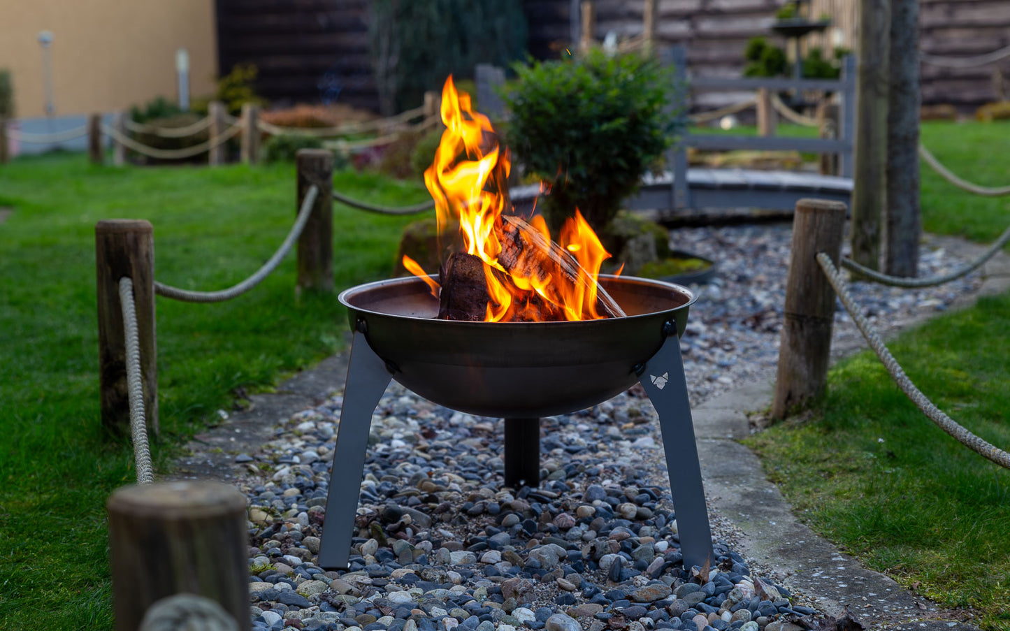 Feuerschale Rustic mit loderndem Feuer auf Kiesbett in einem schön angelegten Garten.