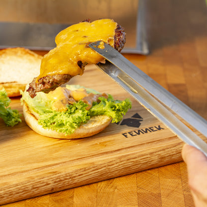 Mit Käse überbackenes Hamburger Patty wird mit Grillzange aus Edelstahl auf ein Brötchen gelegt, darunter ein Holzbrett mit Fennek Logo-Branding.