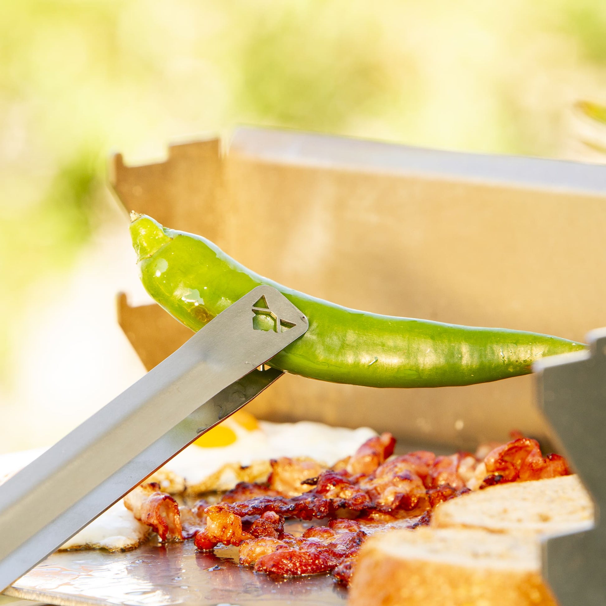 Edelstahl Grillzange mit FENNEK-Logo an der Kontaktfläche herausgeschnitten hält eine Pepperoni von der Plancha.