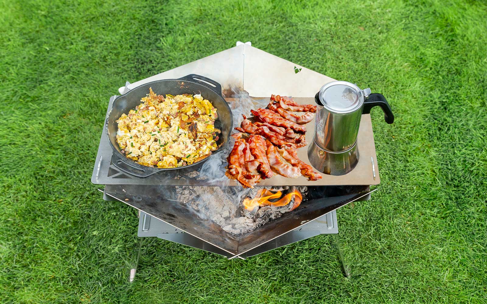 Rührei in der Pfanne, Brötchen, Speck und eine Kaffeekocher auf einer Grillplatte zubereitet die auf einer mobilen Edelstahl-Feuerschale und auf einer Edelstahl-Erhöhung befestigt wurde. Im Hintergrund grüner Rasen.